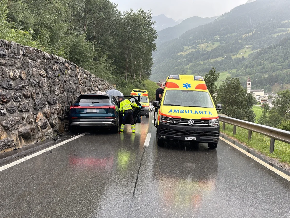 Ein Rettungswagen steht auf einer nassen Bergstraße neben einem beschädigten Auto, das gegen eine Steinmauer gefahren ist. Rettungskräfte sind vor Ort.
