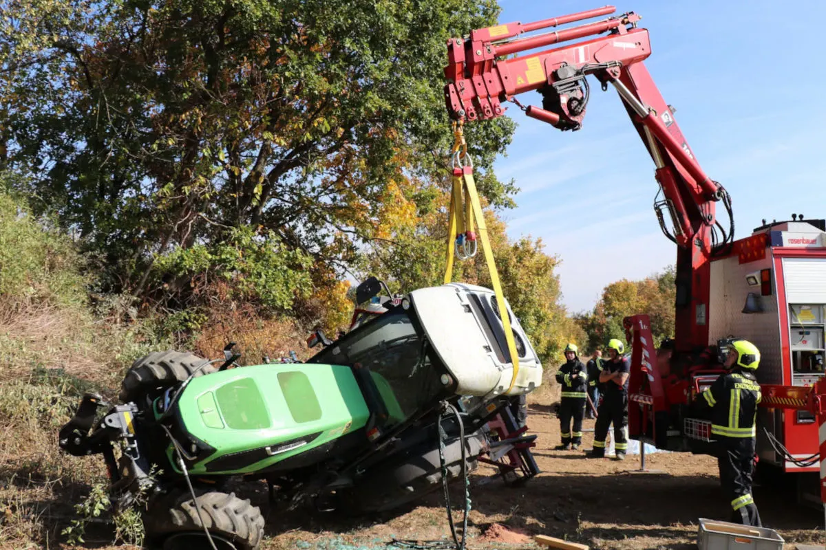 Ein grüner Traktor liegt auf der Seite und wird von einem roten Kran angehoben. Umstehende Feuerwehrleute beobachten die Bergung.