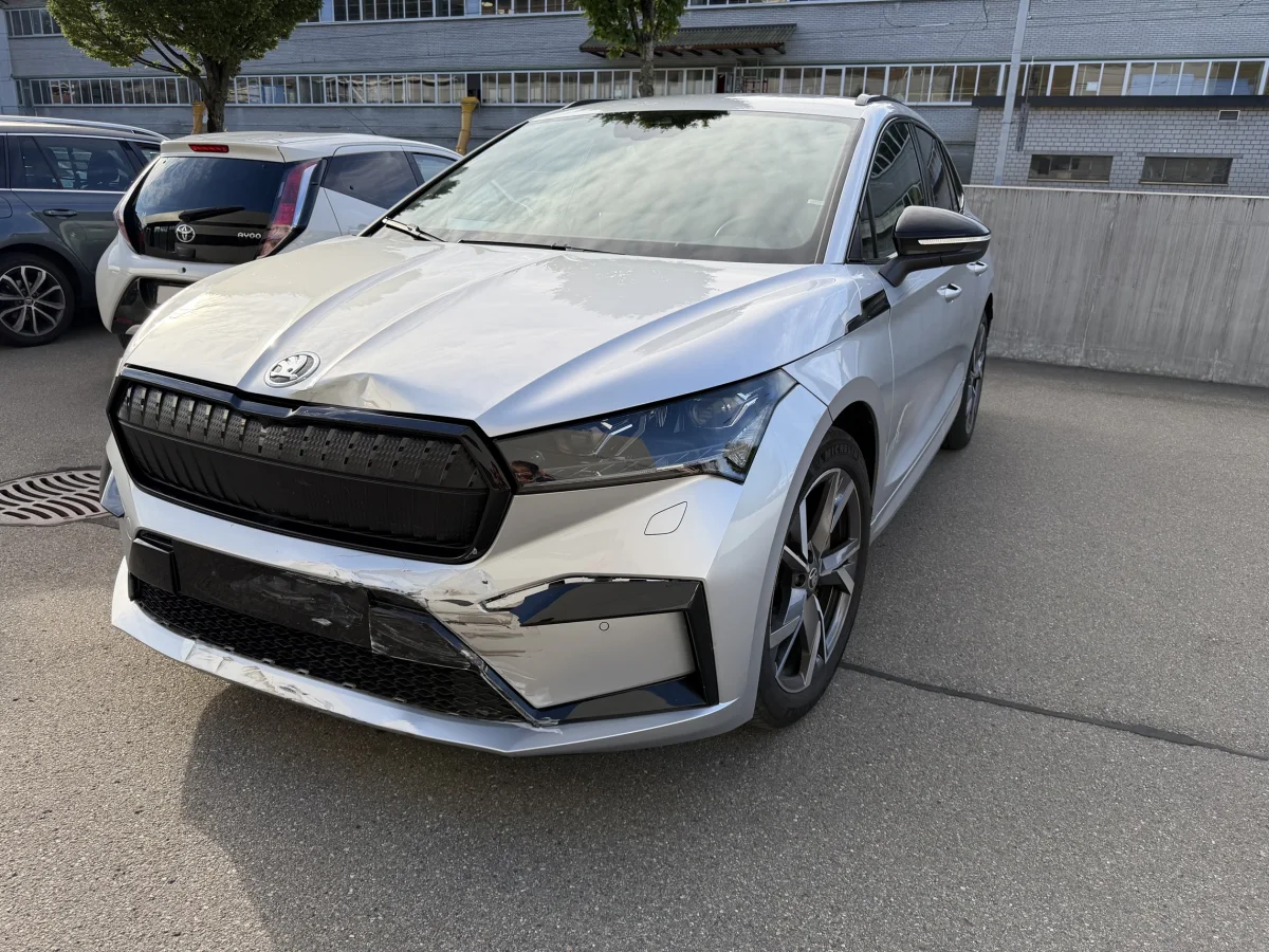 Ein weißer Skoda mit Beschädigungen an der Front steht auf einem Parkplatz. Im Hintergrund sind andere Autos und ein Gebäude sichtbar.