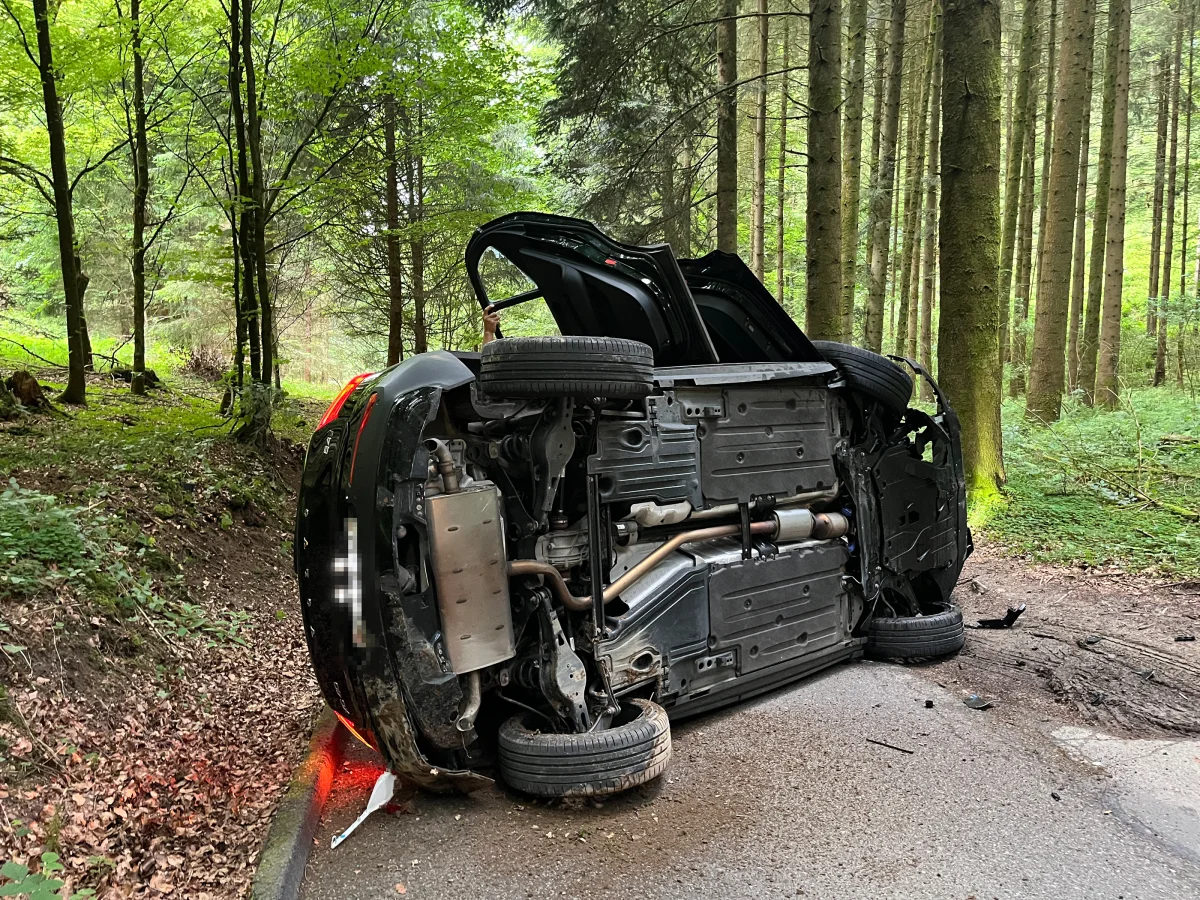 Ein Fahrzeug liegt seitlich auf einer schmalen Straße, umgeben von dichten Bäumen in einem Waldgebiet.