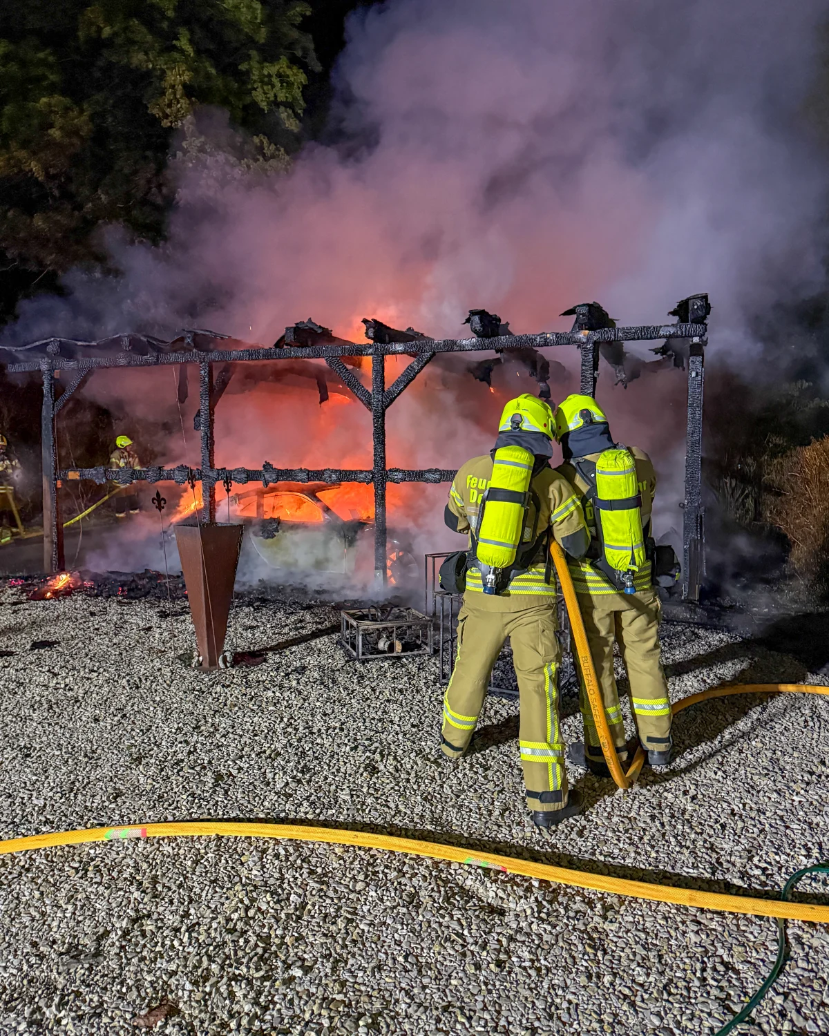 Zwei Feuerwehrleute in voller Einsatzkleidung stehen vor einem brennenden Holzrahmen. Dicker Rauch steigt in die Nachtluft auf.