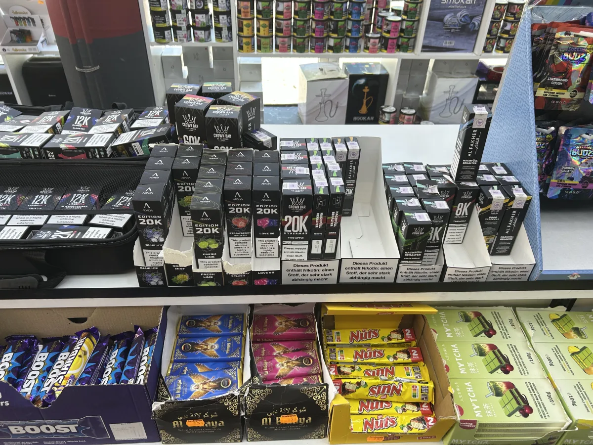 Auf einem Ladentisch sind viele Vaping-Produkte in bunten Verpackungen zusammen mit diversen Süßigkeiten zu sehen. Im Hintergrund stehen Regale mit weiteren Artikeln.