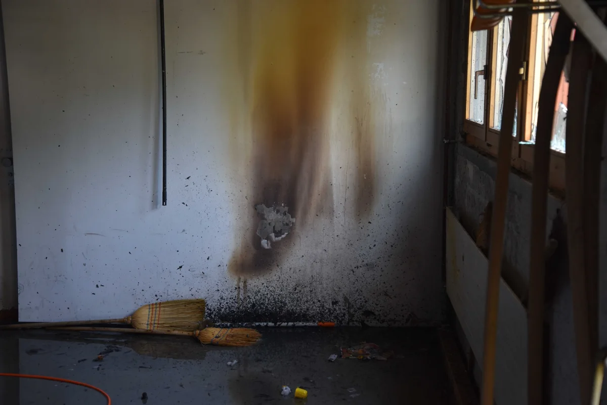 Eine Wand mit starken Rußverfärbungen deutet auf ein Feuer hin. Auf dem Boden liegen ein Besen und verstreuter Müll.