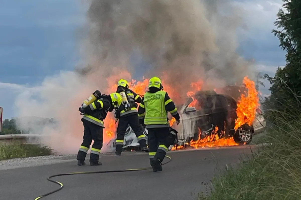 Drei Feuerwehrleute in Schutzkleidung bekämpfen einen brennenden Pkw, umgeben von dichter Rauchentwicklung.