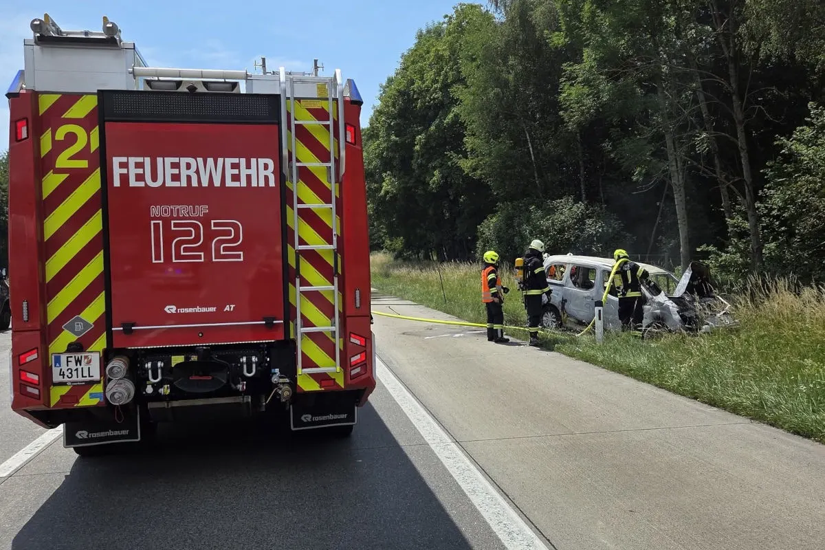 Ein Feuerwehrfahrzeug steht auf der Straße. Im Hintergrund sichern Feuerwehrleute ein ausgebranntes Auto am Straßenrand.