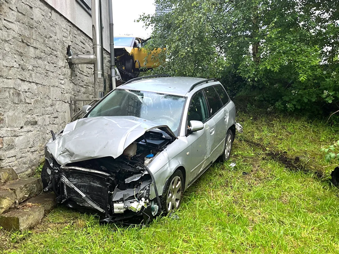 Das Bild zeigt ein stark beschädigtes silbernes Auto, das neben einer Steinmauer auf einer Wiese steht. Die Frontpartie ist eingedrückt, und auf der linken Seite ist ein gelbes Fahrzeug im Hintergrund erkennbar.