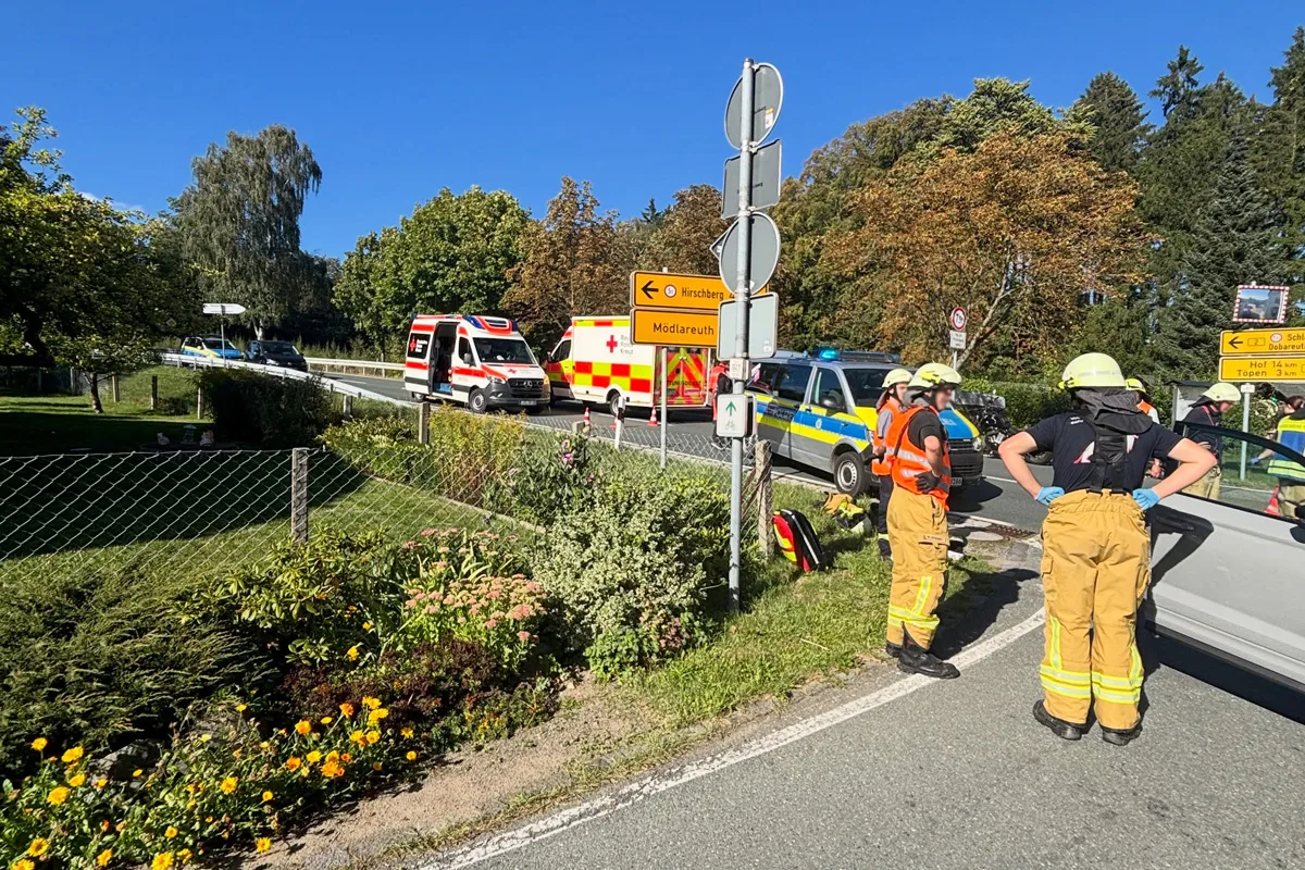 Ein Krankenwagen und ein Polizeiwagen stehen an einer Landstraße, umgeben von Feuerwehrleuten in Einsatzkleidung. Die Szene zeigt eine ländliche Umgebung bei sonnigem Wetter.