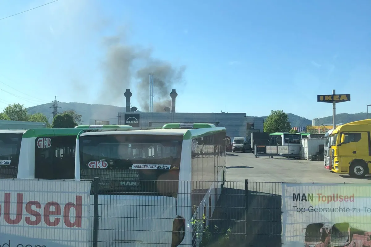 Zwei Busse stehen geparkt auf einem offenen Parkplatz. Im Hintergrund sieht man Rauch, der aus einem Gebäude aufsteigt, in der Ferne ist ein IKEA-Schild sichtbar.