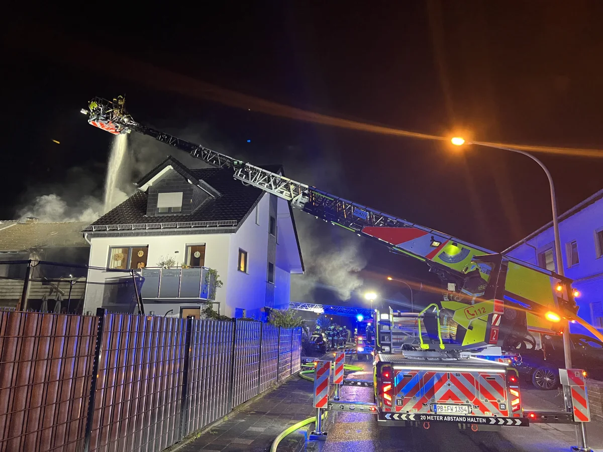 Ein Wohnhaus steht bei Nacht im Flammen. Eine Feuerwehrleiter spritzt Wasser auf das Dach, und Einsatzfahrzeuge stehen vor Ort.
