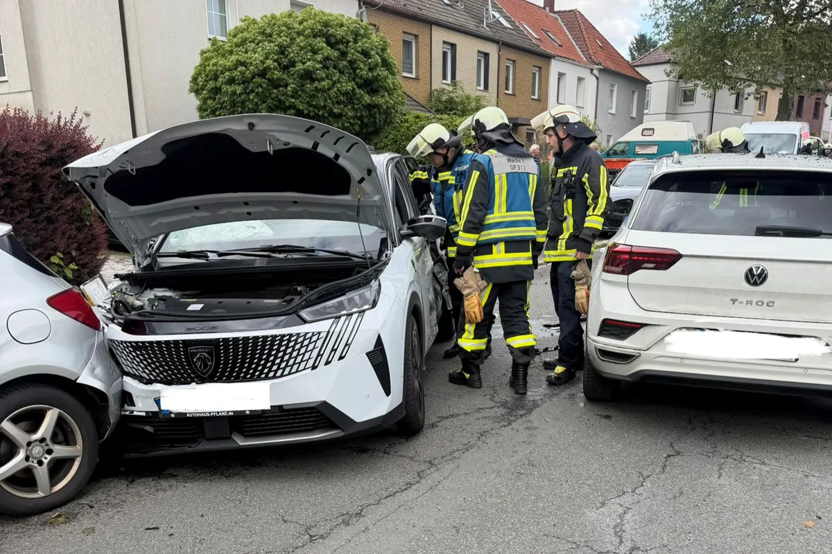Mehrere Feuerwehrleute inspizieren ein beschädigtes, weißes Auto mit geöffneter Motorhaube in einer Wohnstraße. Ein weiteres Auto ist daneben geparkt.
