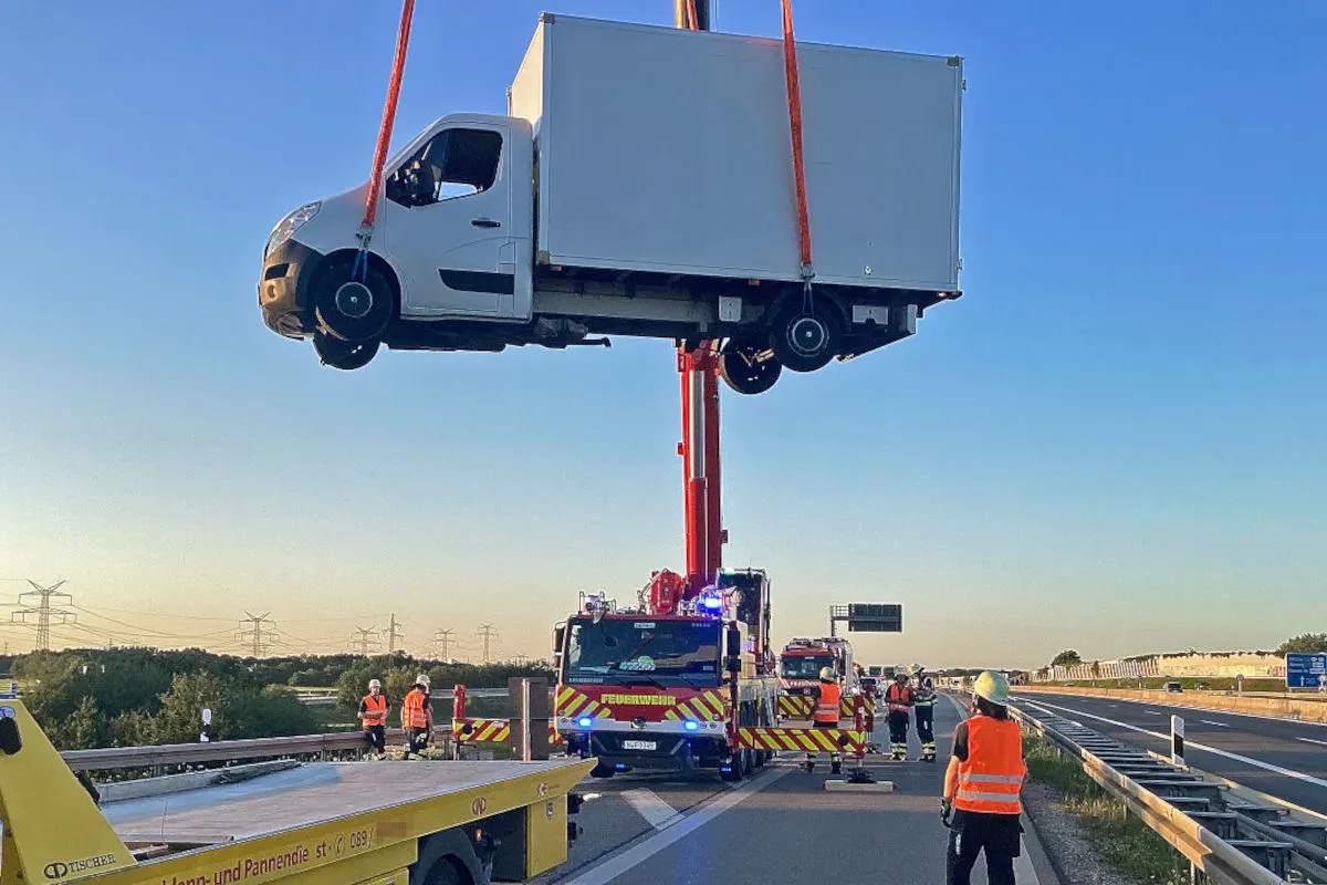 Ein weißer Lieferwagen wird von einem Kran auf einer Autobahn in die Luft gehoben. Feuerwehrleute und Einsatzfahrzeuge sind am Einsatzort sichtbar.