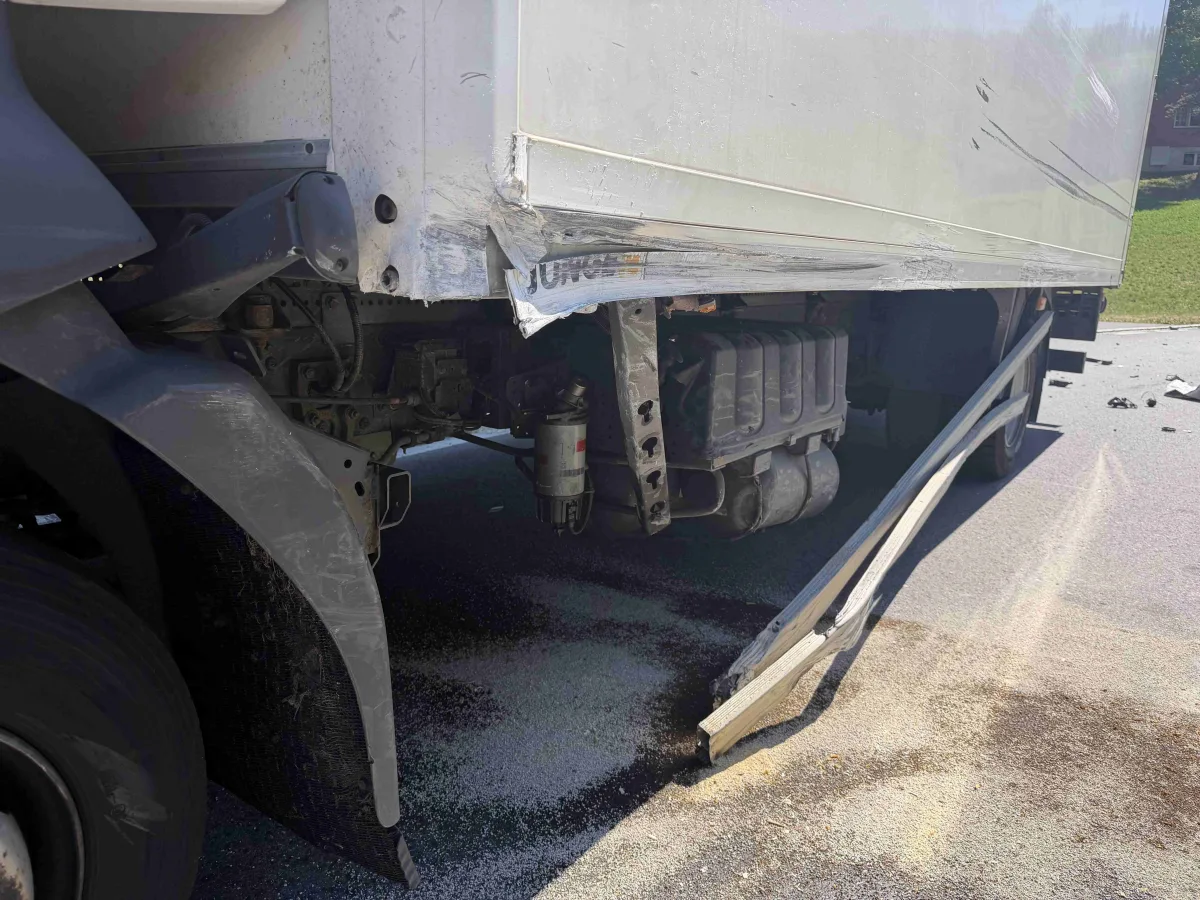 Ein Lastwagen zeigt erhebliche Schäden an der Seite, mit einem verbogenen Metallteil, das auf die Straße hinaussteht. Der Asphalt ist stellenweise mit Schmutz oder Flüssigkeit bedeckt.