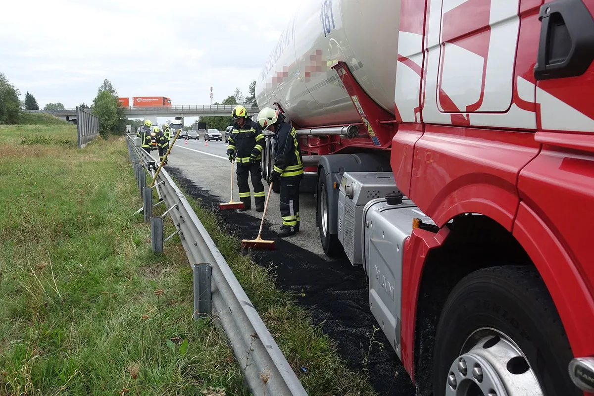 Mehrere Feuerwehrleute stehen neben einem großen roten Tanklastwagen auf einer Autobahn und reinigen die Fahrbahn. Grünflächen flankieren die Straße, während im Hintergrund andere Fahrzeuge vorbeifahren.