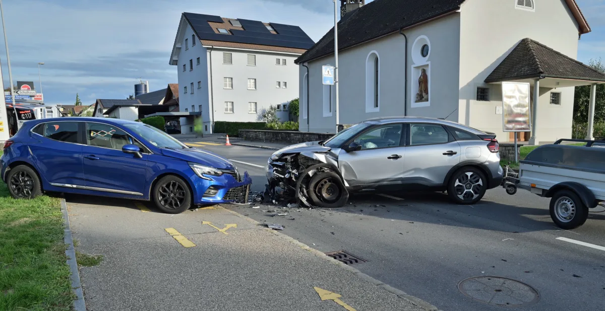 Ein silbernes Auto ist frontal mit einem blauen Auto zusammengestoßen. Trümmerteile liegen auf der Straße, im Hintergrund sind Gebäude zu sehen.