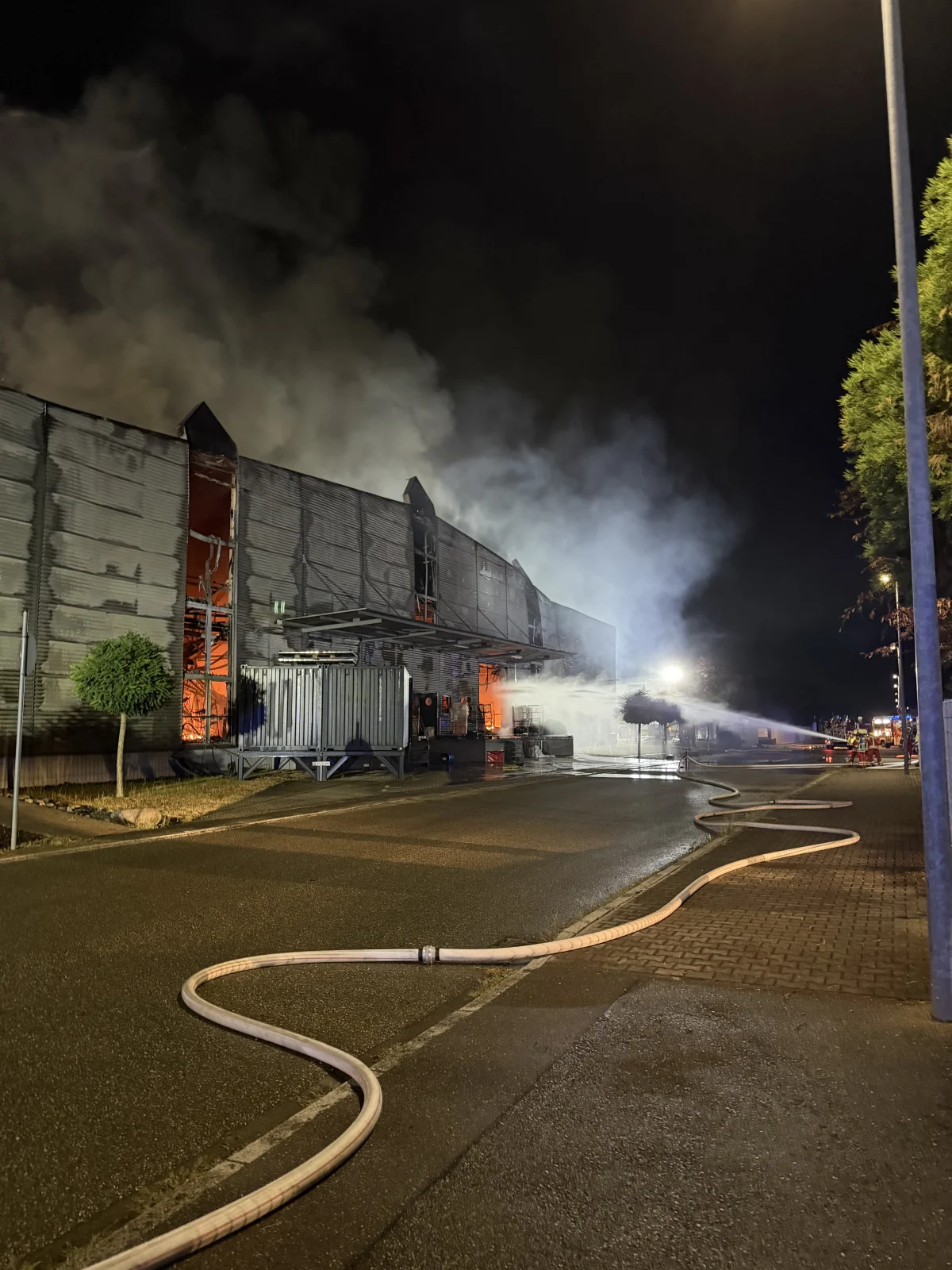 Ein großes Lagergebäude brennt stark; Rauch steigt in die Nachtluft. Feuerwehrschläuche liegen auf der Straße in Richtung des Gebäudes.