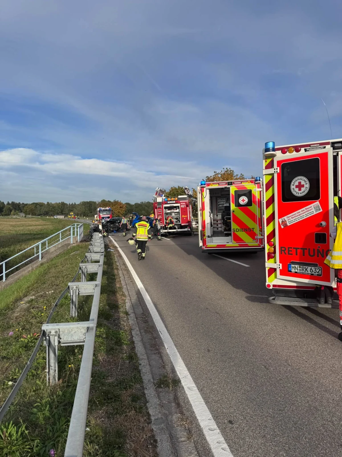 Mehrere Rettungsfahrzeuge sind auf einer Landstraße zu sehen, umgeben von Einsatzkräften in Warnwesten. Der Himmel ist klar mit einigen Wolken und die Szene wirkt herbstlich.