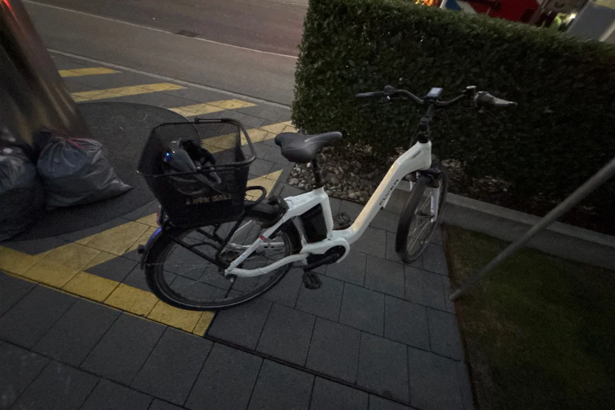 Ein weißes Elektro-Fahrrad mit Korb ist schräg auf einem Gehsteig abgestellt. Im Hintergrund sind Müllsäcke und Pflanzen zu sehen.