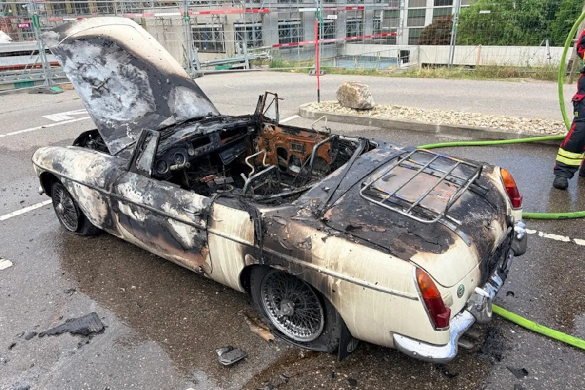 Ein Cabriolet, dessen Lack und Innenraum durch ein Feuer stark beschädigt wurden. Die Motorhaube und der Kofferraum sind geöffnet; ein Feuerwehrschlauch ist am Rand sichtbar.
