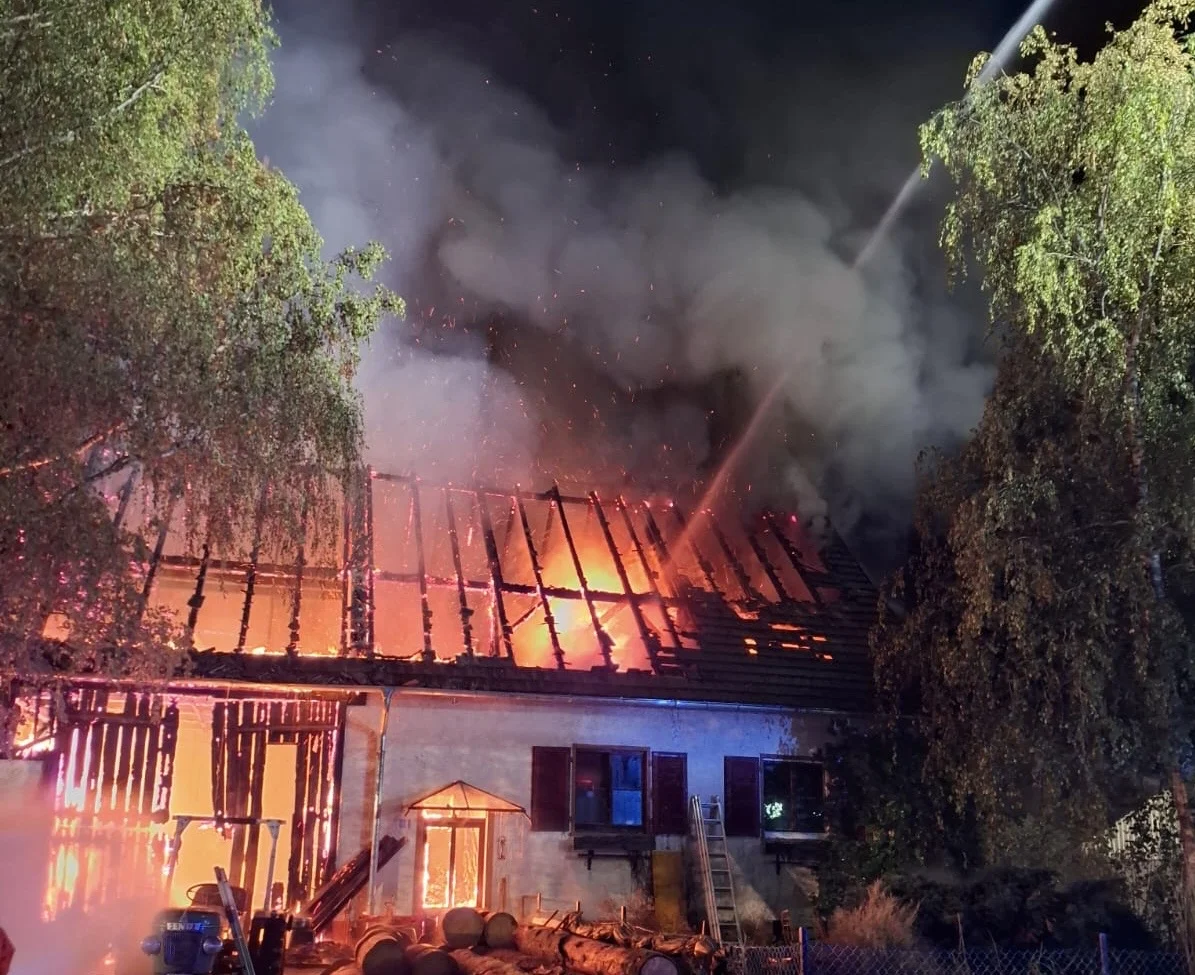 Ein Wohnhaus steht nachts in Flammen, das Dach brennt heftig und Feuerwehrkräfte bekämpfen den Brand mit Wasserstrahlen.