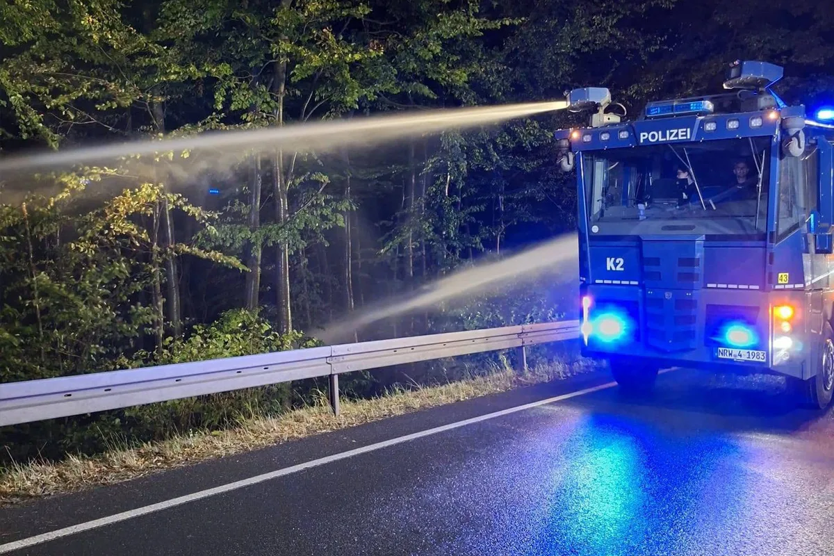 Ein blaues Polizeifahrzeug sprüht Wasser auf eine nasse Landstraße neben einem Wald in der Dunkelheit.