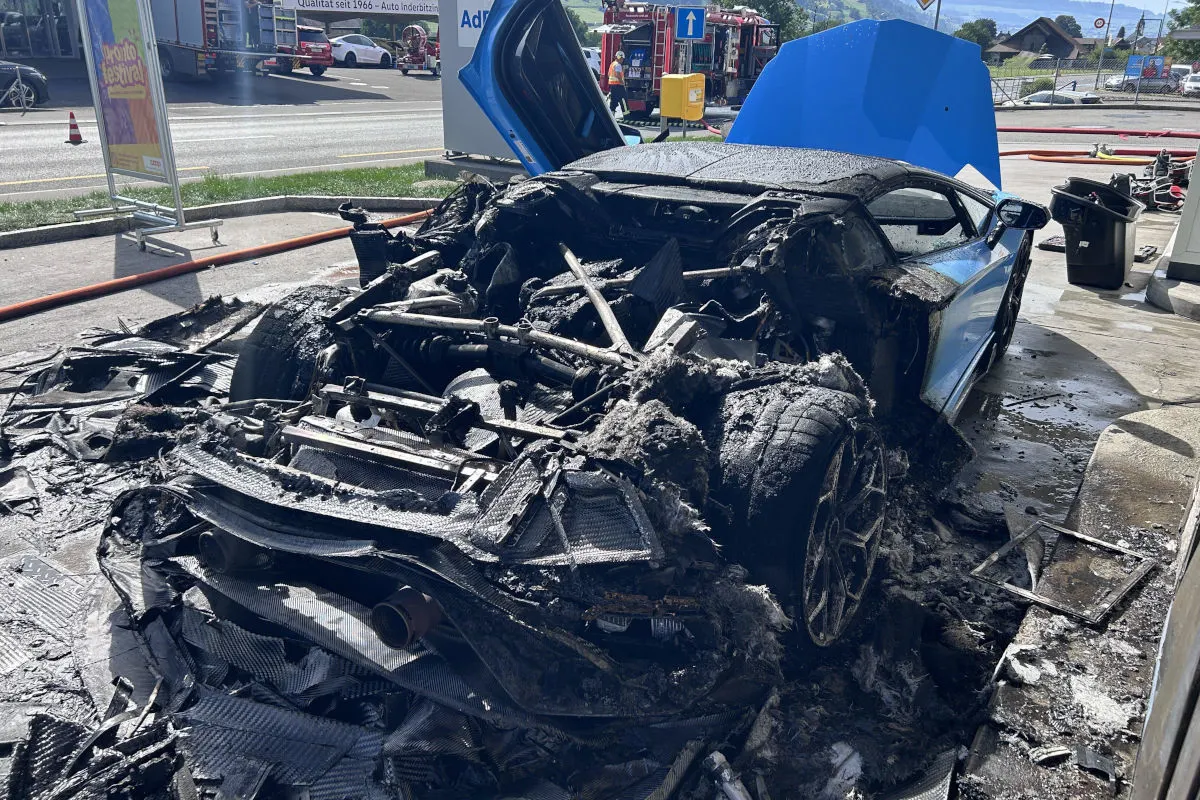 Ein ausgebranntes Fahrzeug steht inmitten von Feuerwehrschläuchen und Trümmern. Im Hintergrund sind Feuerwehrautos zu sehen.