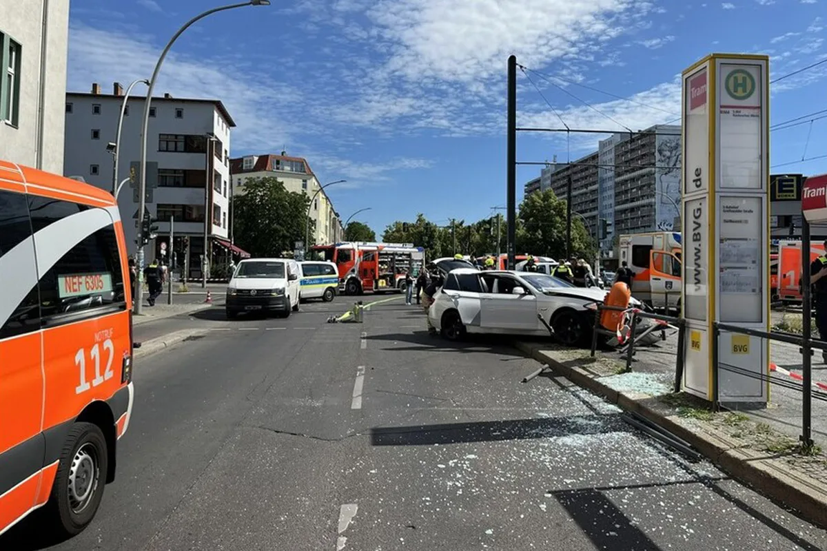 Ein weißes Auto mit Schäden steht nahe einer Berliner Haltestelle, umgeben von Einsatzfahrzeugen bei sonnigem Wetter.