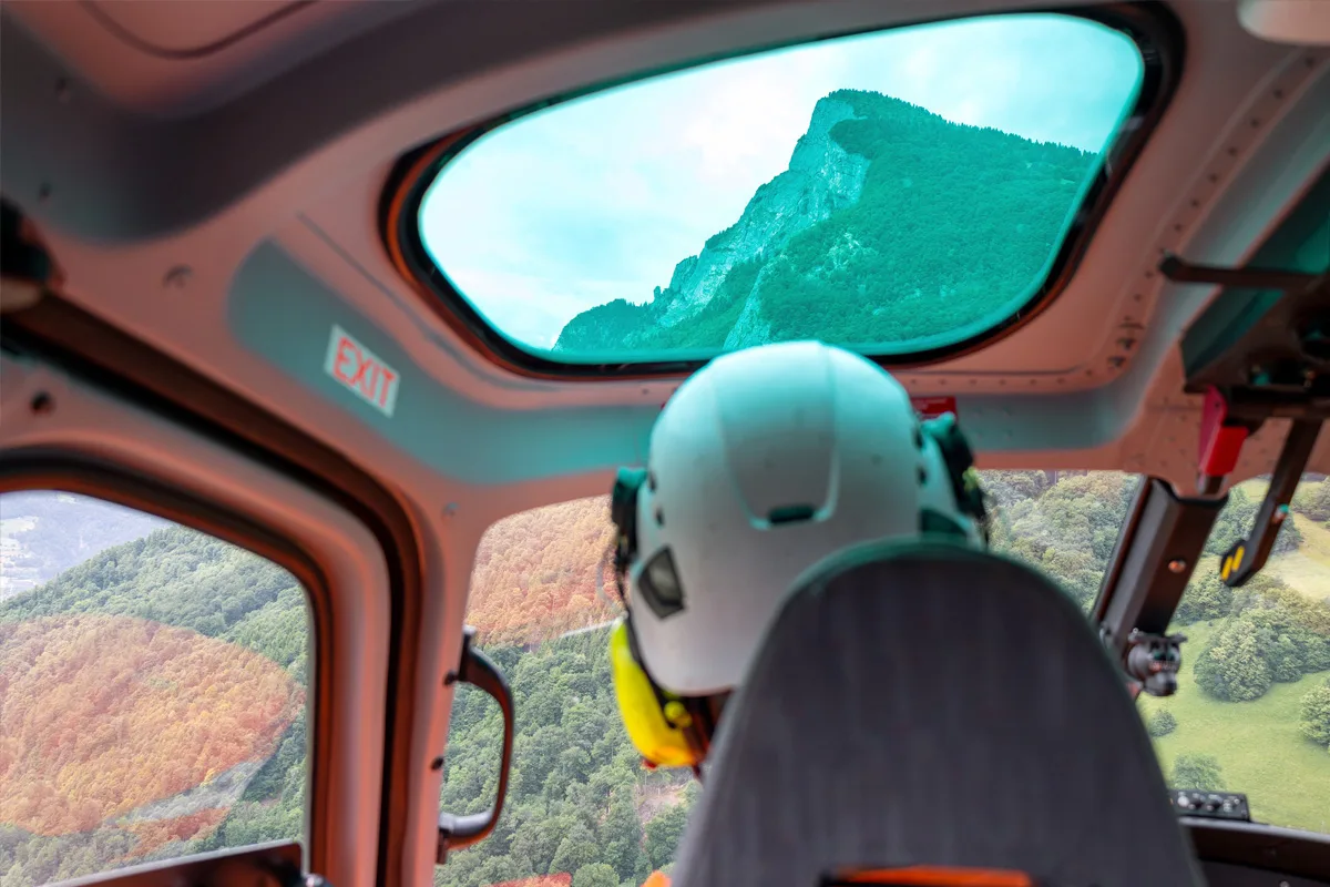 Ein Pilot mit Helm sitzt im Cockpit eines Hubschraubers, während dieser über eine grüne, bergige Landschaft fliegt.