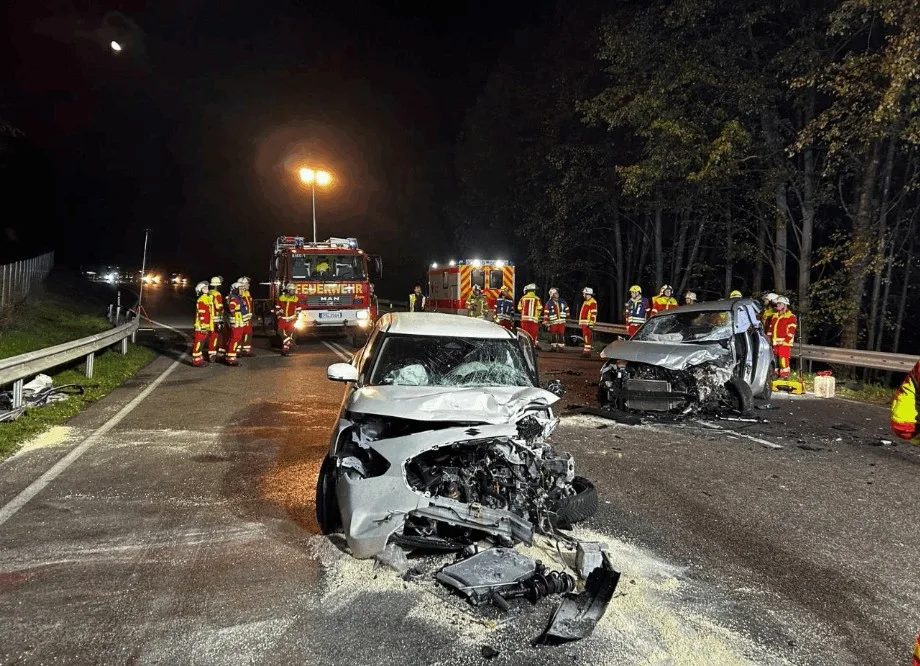 Auf einer nächtlichen Straße sind zwei stark beschädigte Fahrzeuge nach einer Kollision zu sehen. Im Hintergrund ist die Feuerwehr bei den Sicherungsarbeiten zu erkennen.
