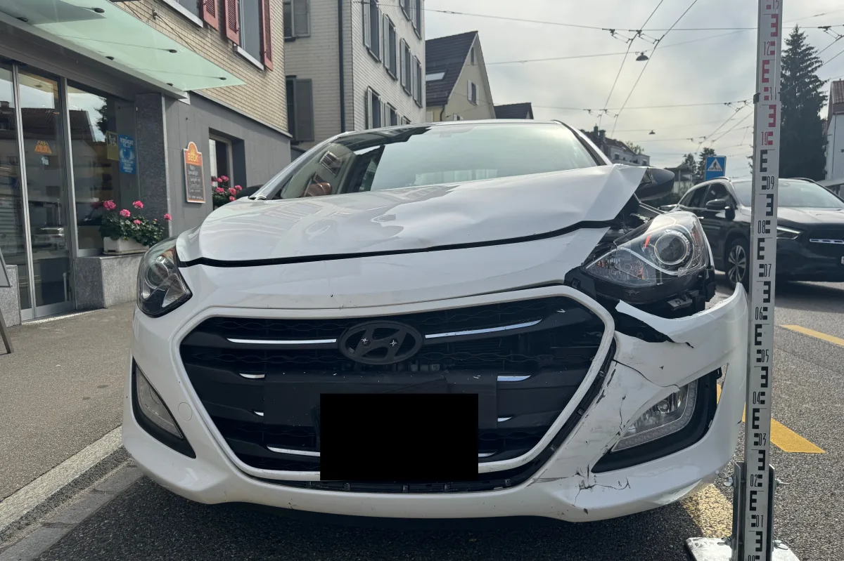 Ein weißer Hyundai mit stark beschädigter Front steht auf einer Stadtstraße vor einem Gebäude. Daneben befindet sich ein Höhenmesspfosten.