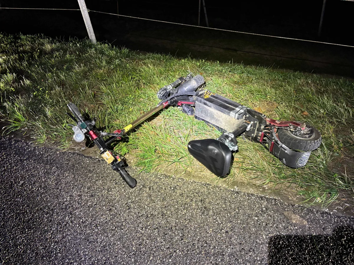 Ein beschädigter E-Scooter liegt auf der Seite in einer grasbewachsenen Fläche neben einer asphaltierten Straße. Es ist Nacht.