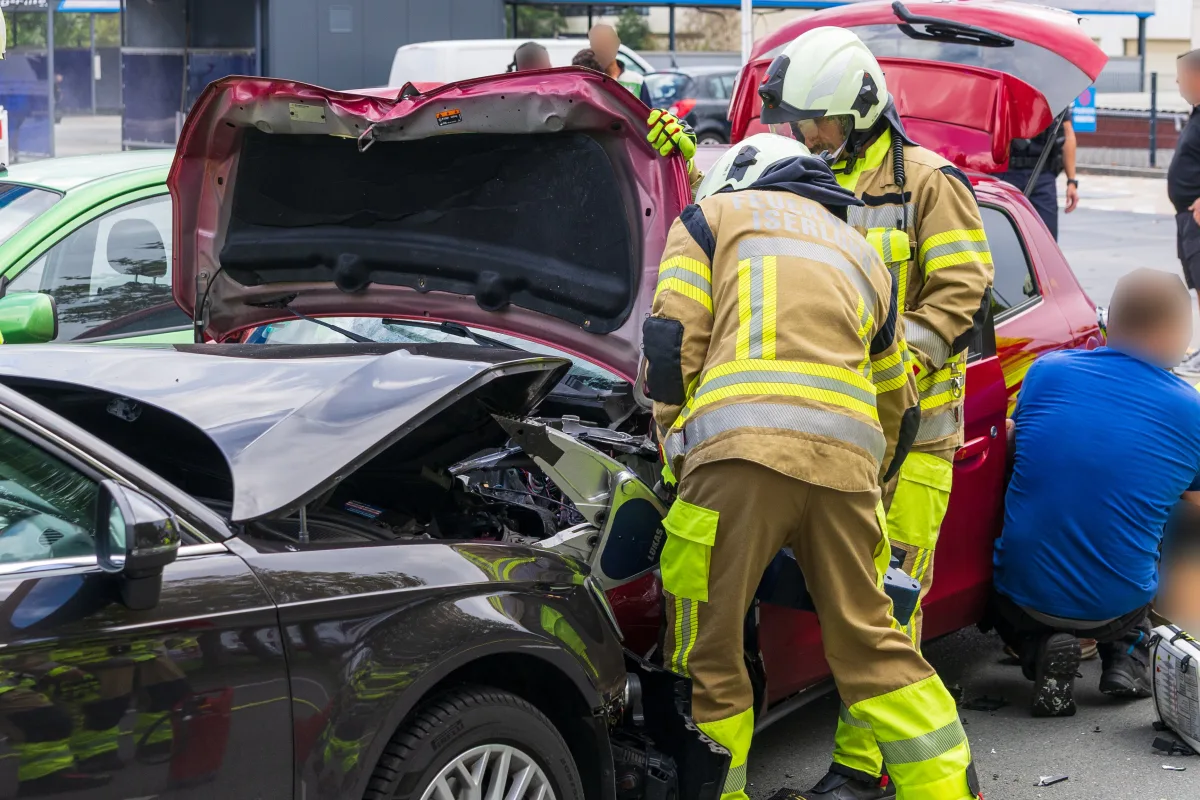 Zwei Feuerwehrleute in Schutzkleidung stehen an einem grauen Auto, dessen Motorhaube geöffnet ist. Ein rotes Auto im Hintergrund.
