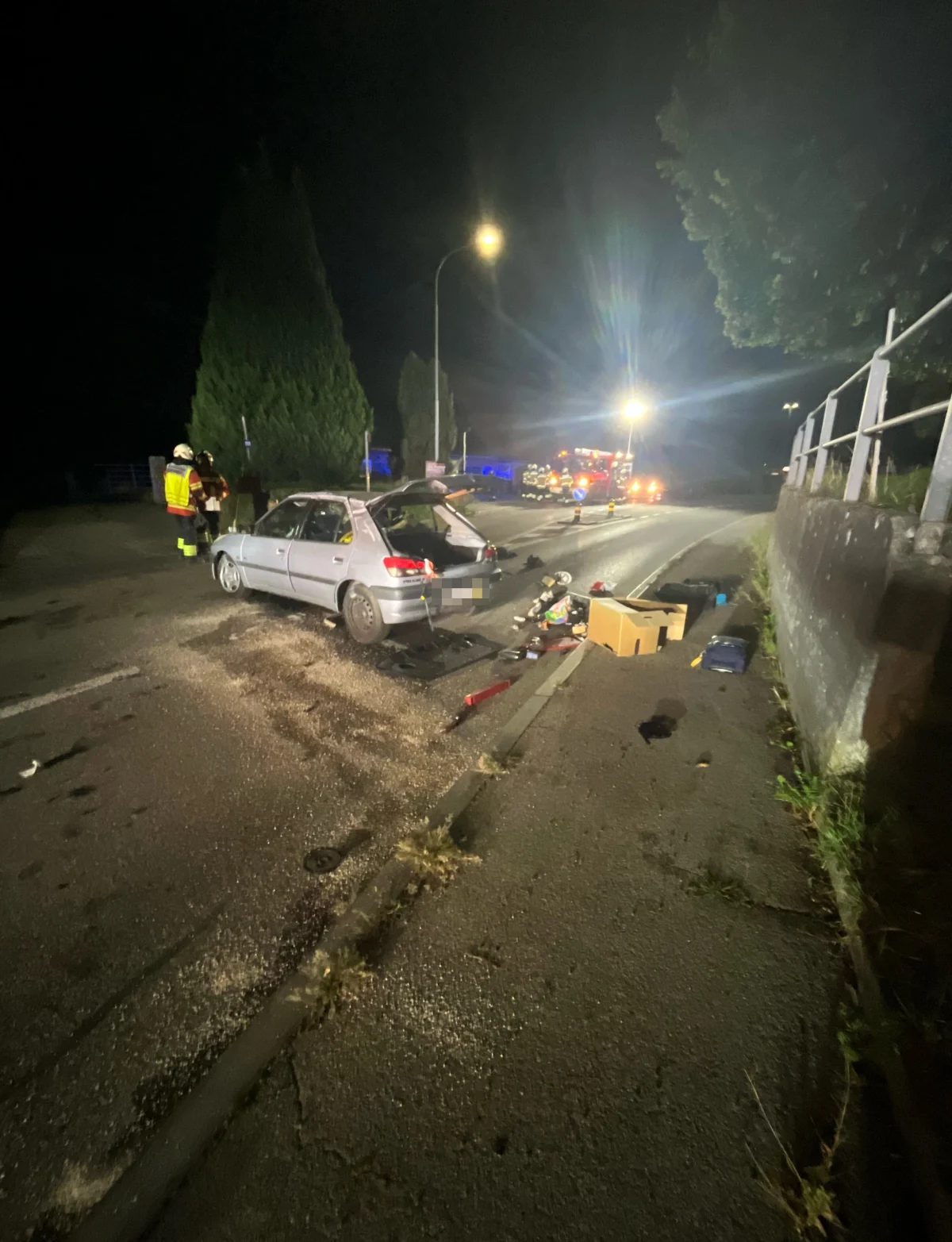 Ein silbernes Auto steht mit geöffnetem Kofferraum auf einer nächtlichen Straße. Feuerwehrleute sind vor Ort, kein Feuer sichtbar.