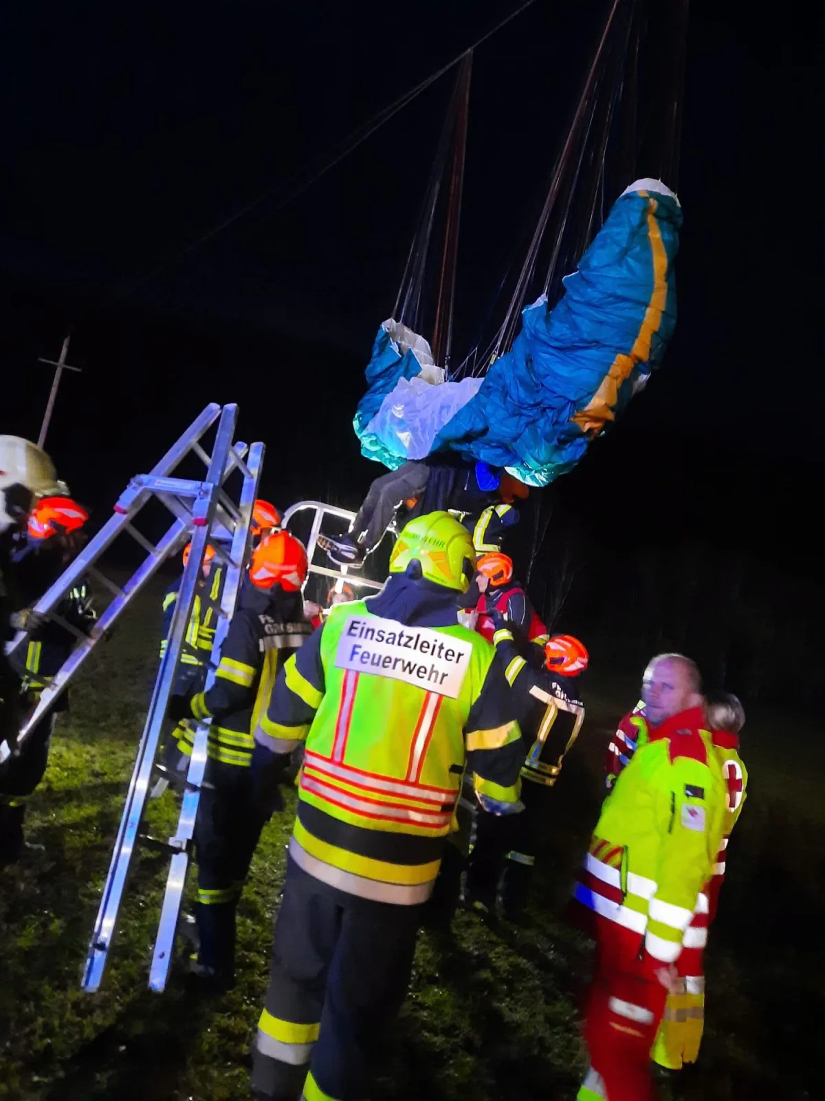 Mehrere Feuerwehrleute arbeiten nachts mit einer Leiter, um einen verletzten Paraglider zu bergen, der in einer Höhe hängt. Notfallausrüstung ist im Einsatz.