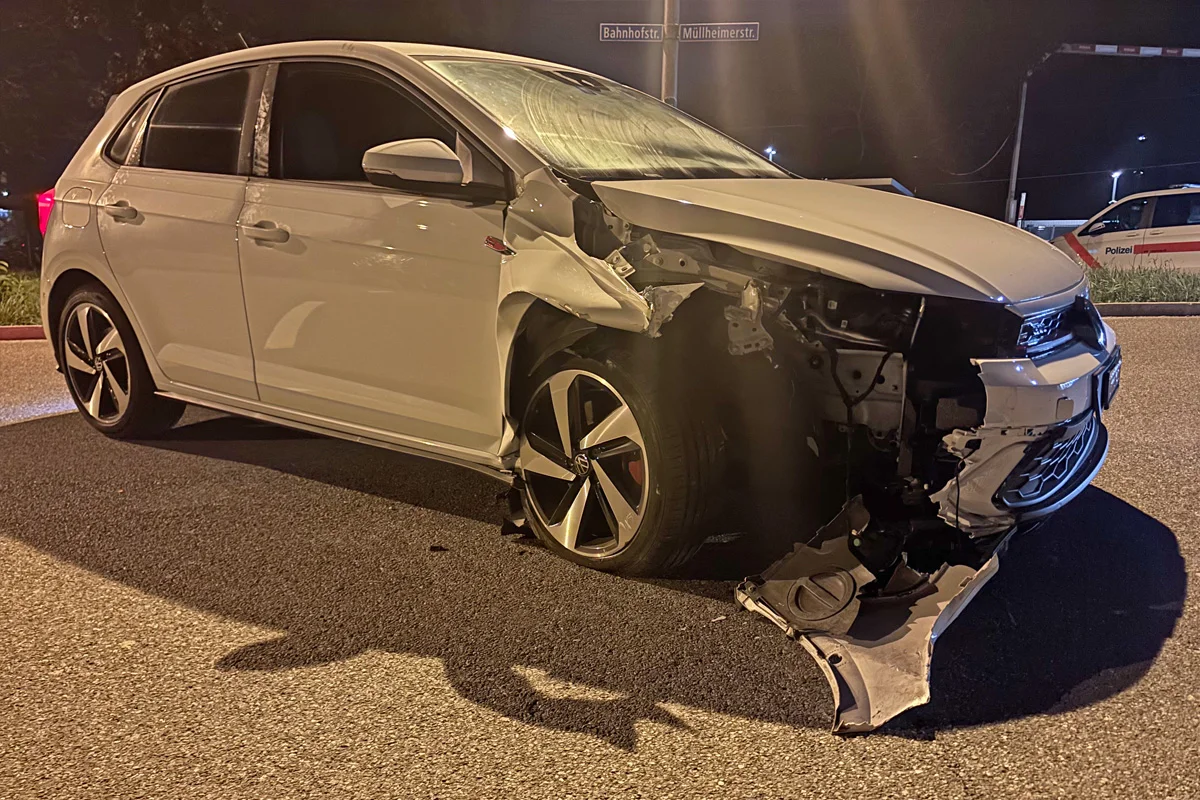 Ein weißer VW steht auf einer Straße mit der stark beschädigten Front. Im Hintergrund sind Straßenschilder und ein Polizeifahrzeug sichtbar.