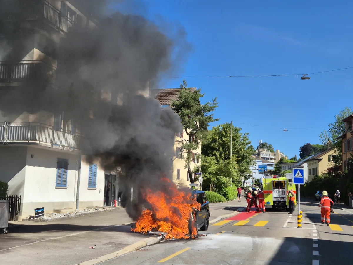 Ein Auto steht im Vollbrand vor einem Wohngebäude, schwarzer Rauch steigt auf; Feuerwehrleute löschen das Feuer.