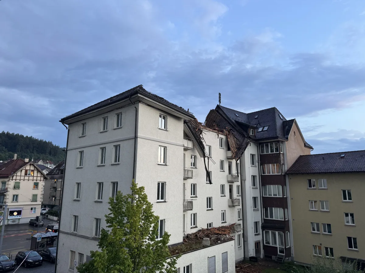 Das Bild zeigt das stark beschädigte Dach eines Mehrfamilienhauses nach einem Sturm. Teile des Daches sind eingestürzt und Trümmer liegen sichtbar auf dem Boden.