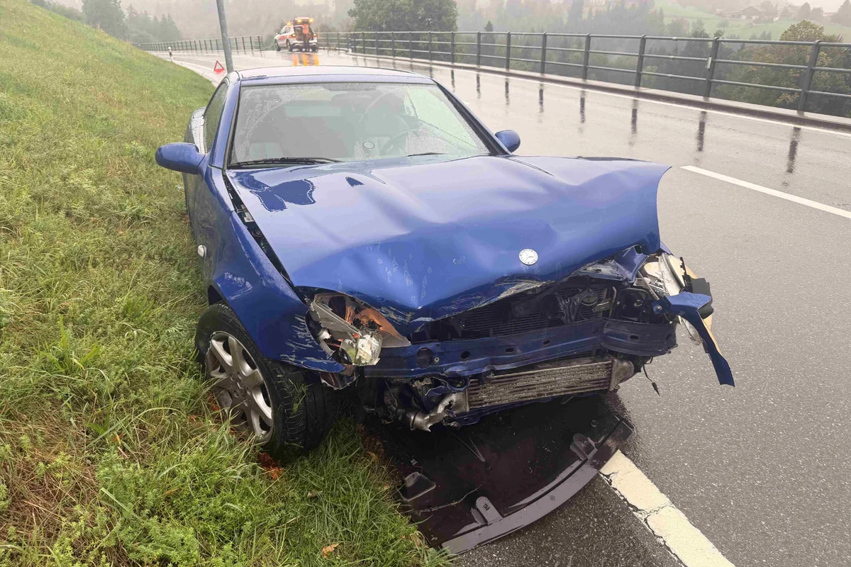 Ein blauer Pkw steht beschädigt am Straßenrand einer nassen Fahrbahn. Im Hintergrund ist ein Abschleppwagen zu sehen.
