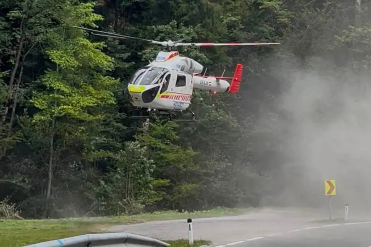 Ein Rettungshubschrauber mit markanten roten und gelben Markierungen schwebt über einer Straße in einem bewaldeten Gebiet. Es herrscht Tageslicht und der Hubschrauber scheint im Begriff zu landen.