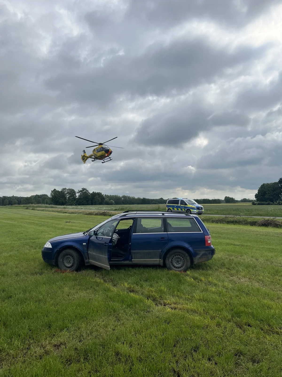 Ein blauer Pkw steht mit offener Autotür auf einer Wiese. Im Hintergrund ist ein Rettungshubschrauber im Anflug zu sehen.