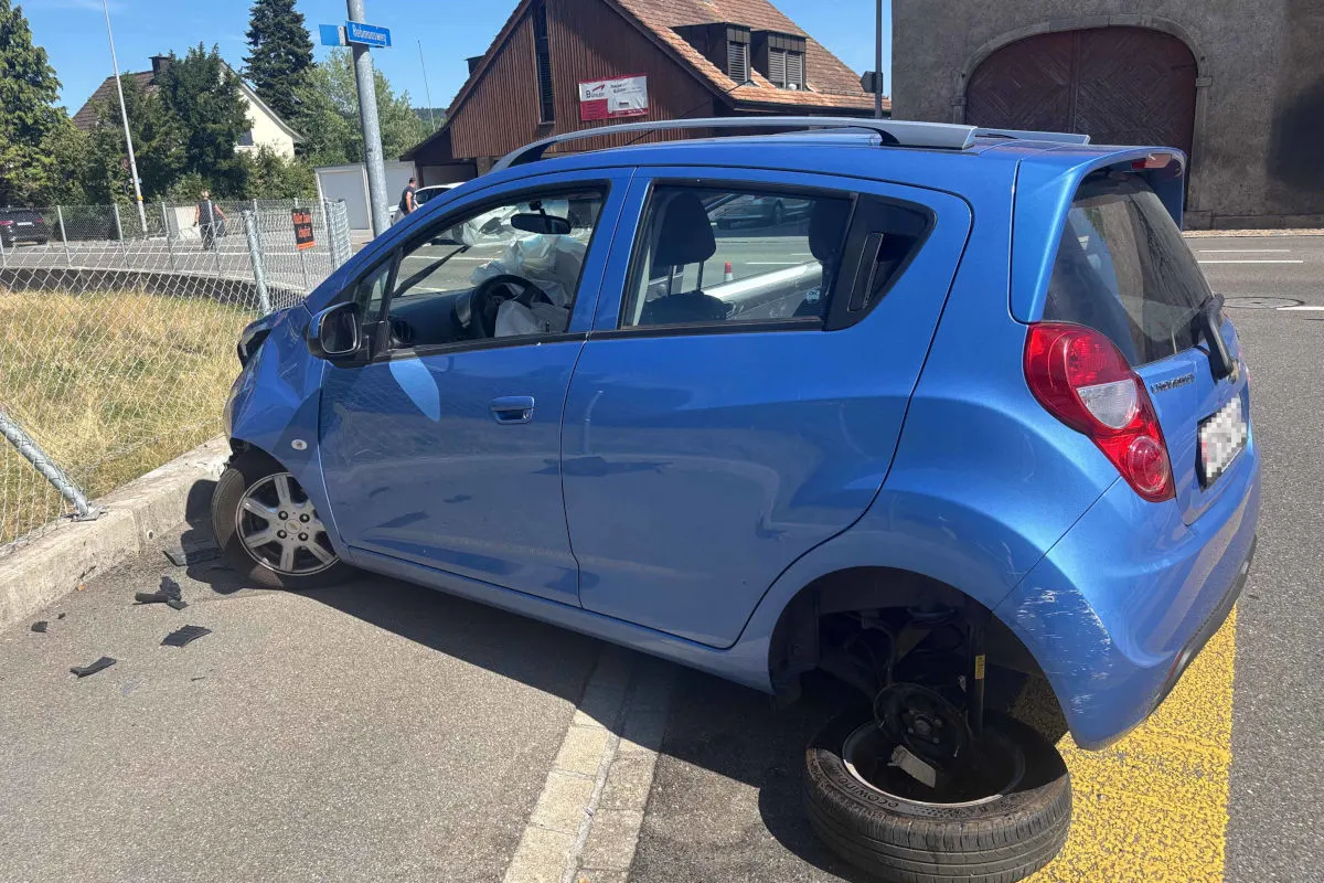Ein blauer Kleinwagen steht am Straßenrand, der Vorderreifen auf der Fahrerseite ist abgerissen. Im Hintergrund sind ein Holzzaun und Wohnhäuser zu sehen.