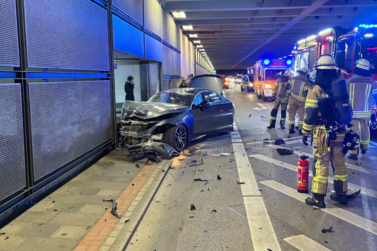 Ein stark zerstörtes Auto steht frontal an einer Wand mit geöffneter Motorhaube und umgeben von Feuerwehrleuten in Ausrüstung.