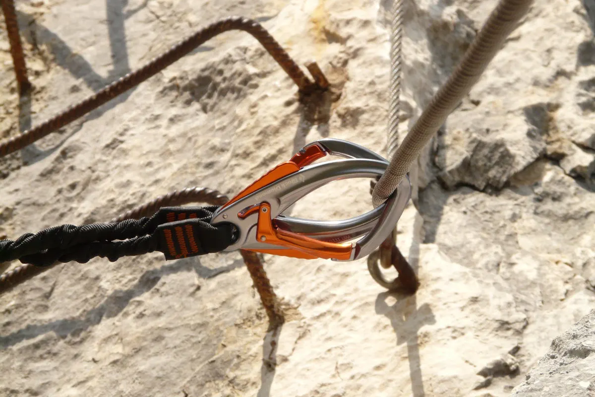 Ein orange-grauer Kletterkarabiner ist an einem Stahlseil befestigt, das an einem Felsen verankert ist. Der Karabiner zeigt typische Merkmale eines Klettersteigsets.