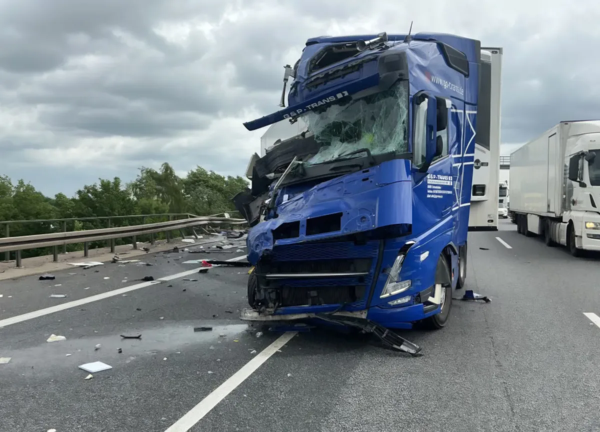 Ein blauer Lkw mit stark beschädigter Vorderseite steht quer zur Fahrbahn auf der Autobahn. Um ihn sind Trümmer verteilt, und weitere Lkw befinden sich auf der Straße.