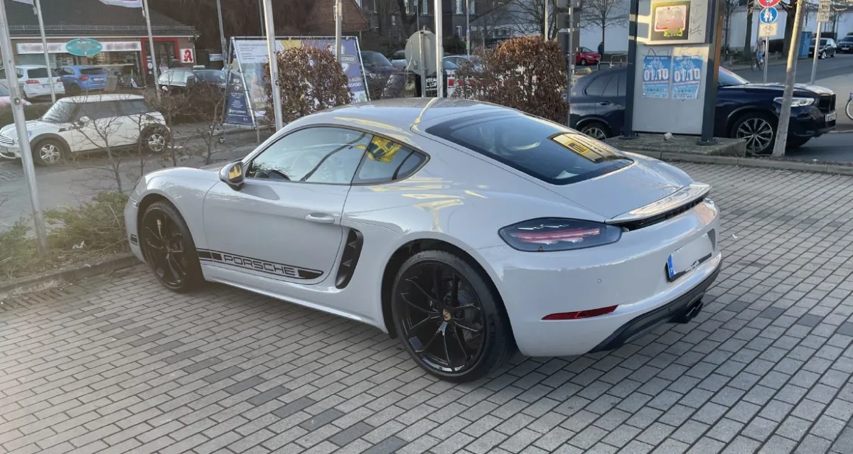 Ein weißer Porsche ist auf einem gepflasterten Parkplatz vor mehreren Geschäften abgestellt. Im Hintergrund sind weitere Fahrzeuge und Werbeschilder zu sehen.