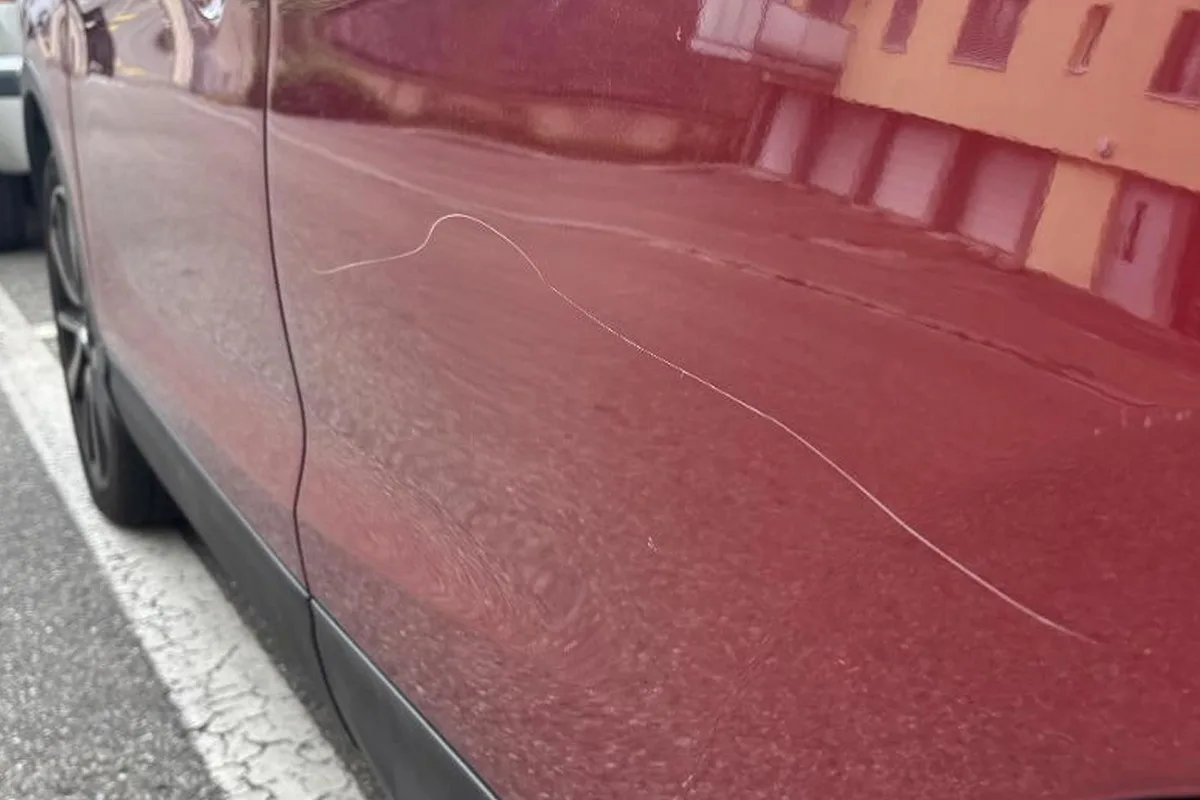 Nahaufnahme einer langen, wellenförmigen Kratzspur auf der Beifahrerseite eines roten Autos auf einem Parkplatz.