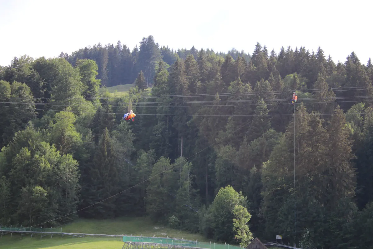 Zwei Personen in orangener Schutzkleidung sind an Seilen über einer dichten Waldlandschaft angebracht. Sie scheinen an einer Hochspannungsleitung zu arbeiten.
