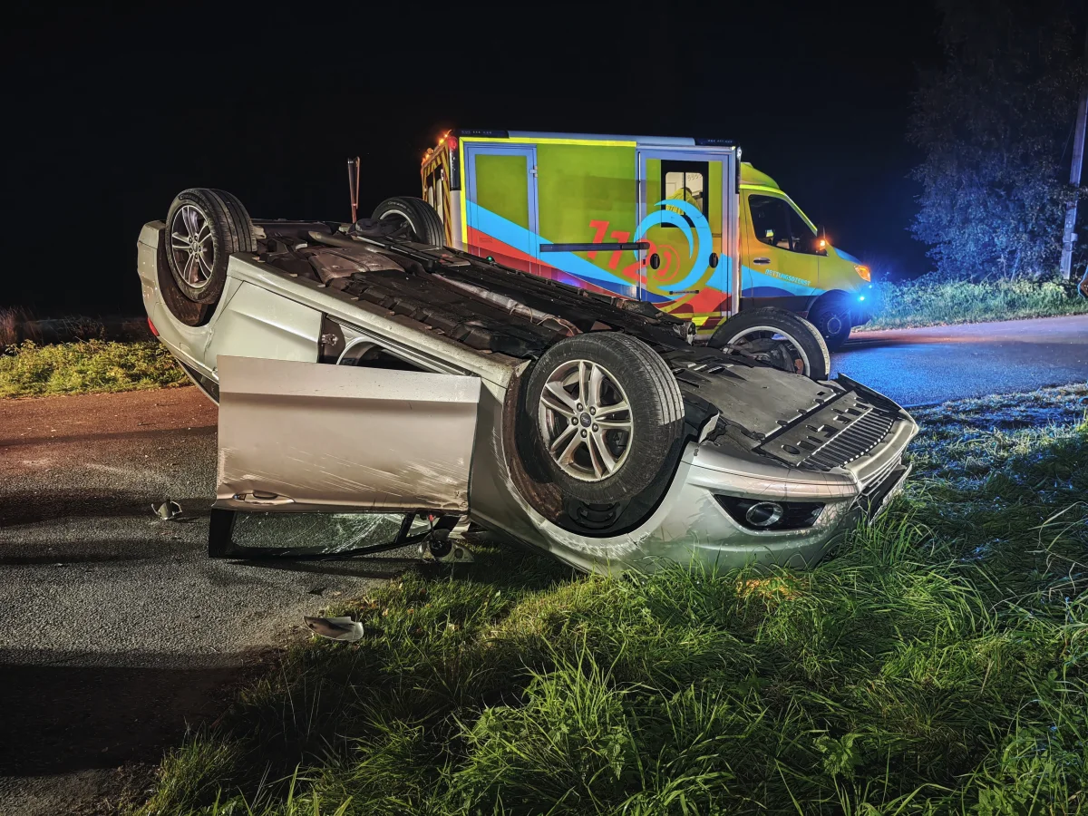 Ein Auto liegt auf dem Dach auf einer Straße, die Fahrertür ist geöffnet. Im Hintergrund ist ein Rettungsdienstfahrzeug mit blinkenden Lichtern sichtbar.