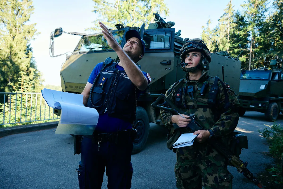 Ein Polizist in Uniform und ein Soldat in Tarnkleidung stehen neben einem gepanzerten Militärfahrzeug auf einer Straße und blicken in dieselbe Richtung.