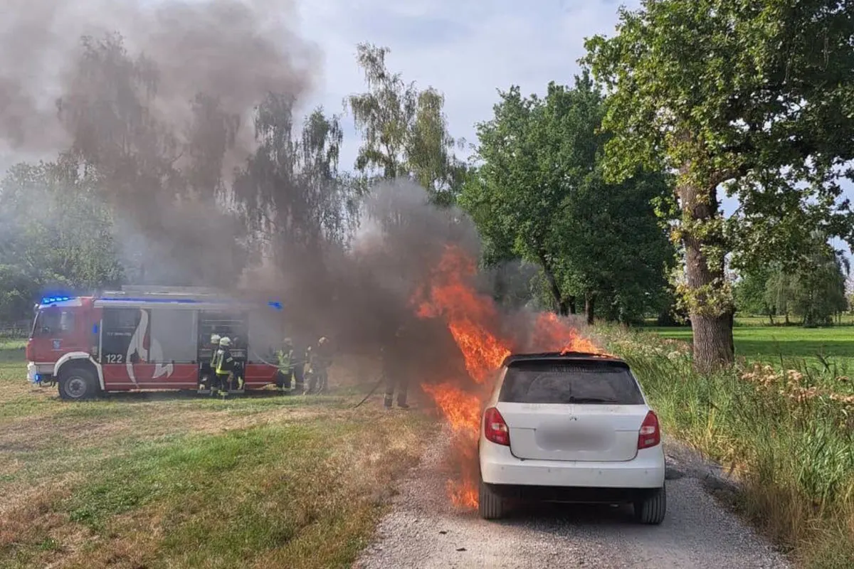 Ein weißes Auto steht am Straßenrand in Flammen, während Feuerwehrleute in der Nähe mit Löscharbeiten beschäftigt sind. Im Hintergrund ist ein rotes Feuerwehrfahrzeug zu sehen.