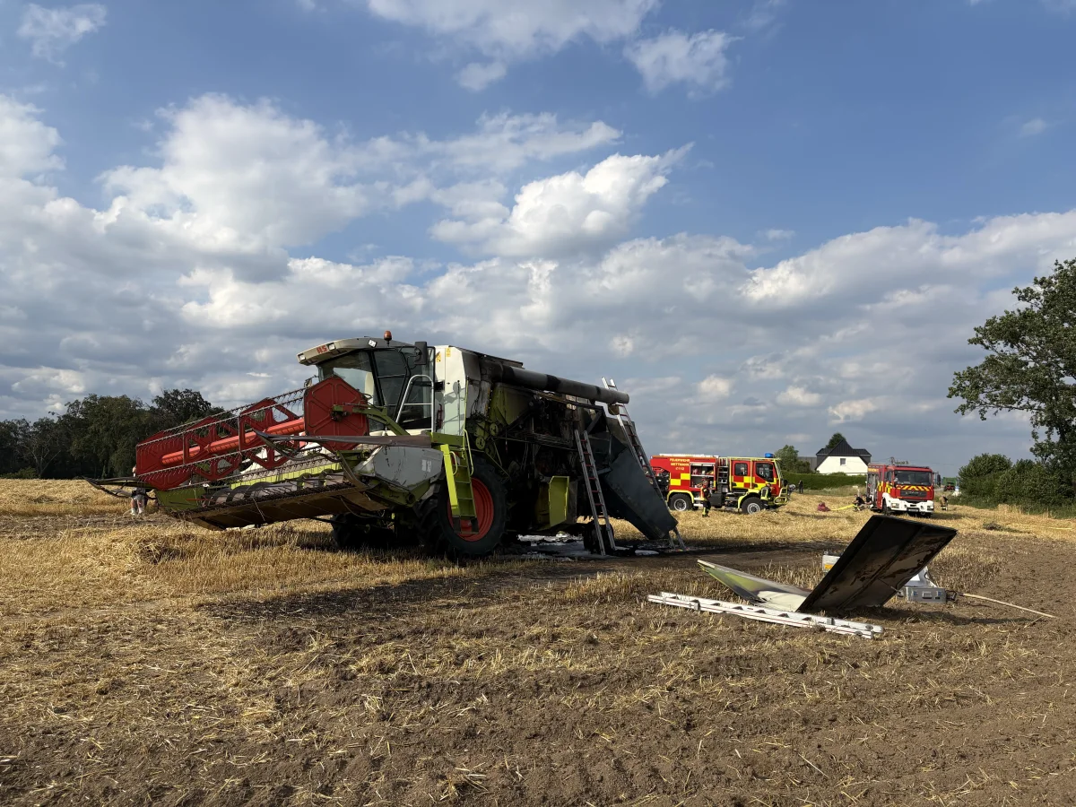 Auf einem abgeernteten Feld steht ein beschädigter Mähdrescher. Feuerwehrfahrzeuge sind im Hintergrund zu sehen.